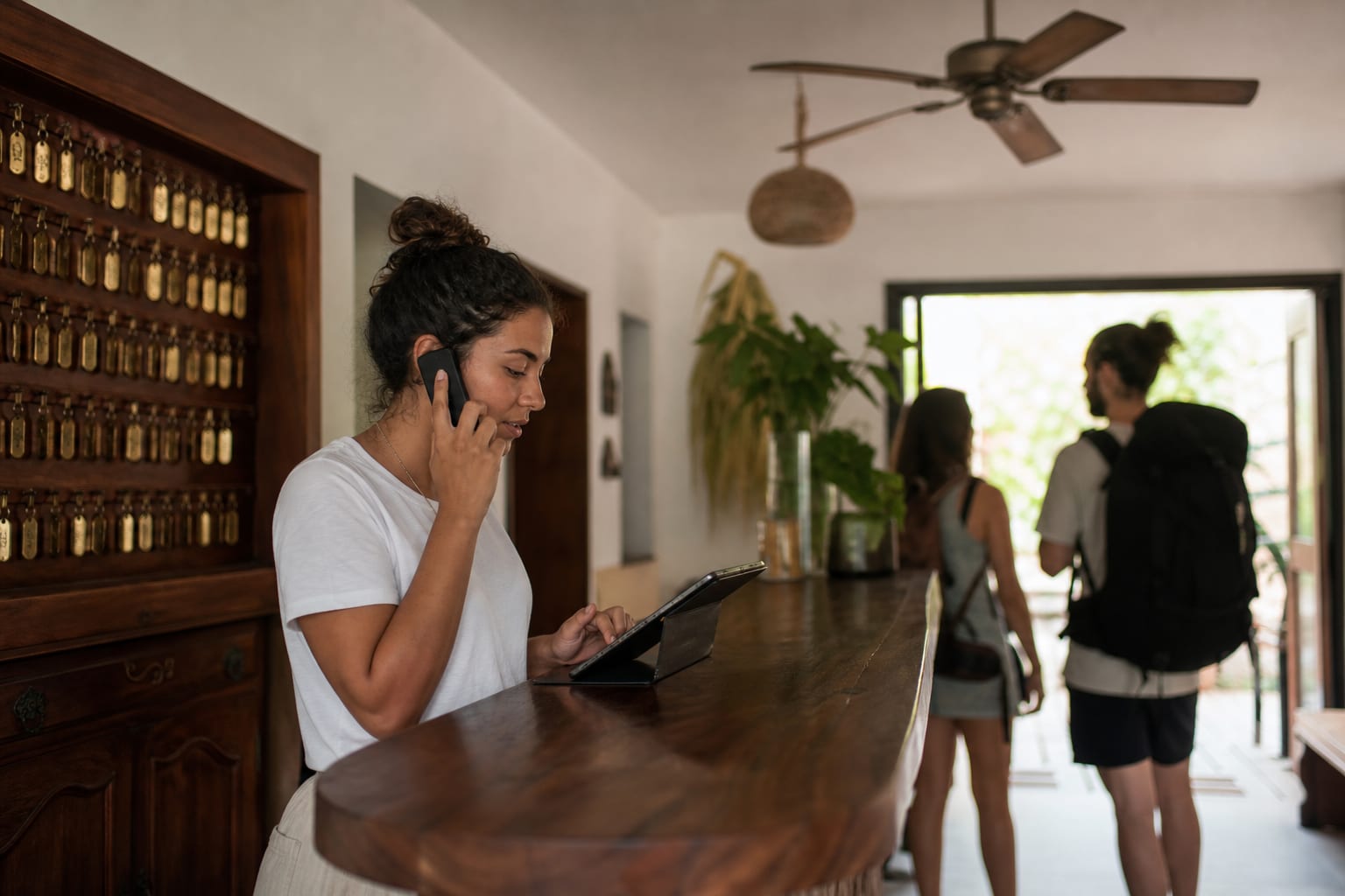 Recepcionista de pousada atende pelo celular com tablet no balcão e quadro de chaves ao fundo